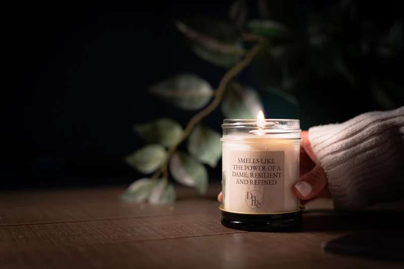 A hand in a cream sweater holds a lit Dame Renovations candle on a wooden table against softly blurred dark foliage.