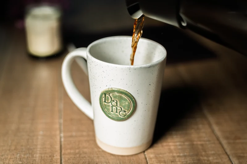 Coffee pours into a tall speckled Dame Renovations mug on a wooden table, with a candle softly blurred behind it.