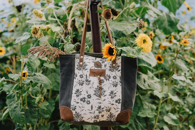 A floral-patterned tote with brown handles and a sunflower hangs among green leaves and yellow flowers.