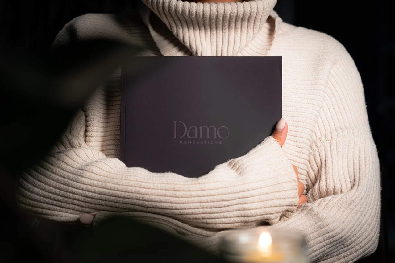 A person in a cream knit sweater holds a closed dark Dame Renovations journal against their chest, with a softly blurred candle in the foreground.