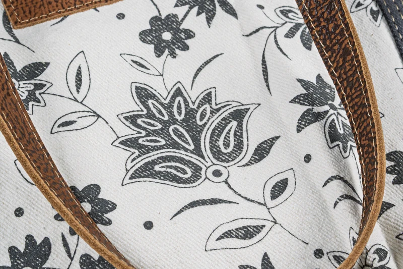 Close view of a black floral pattern on cream tote fabric, framed by textured brown straps and stitching.