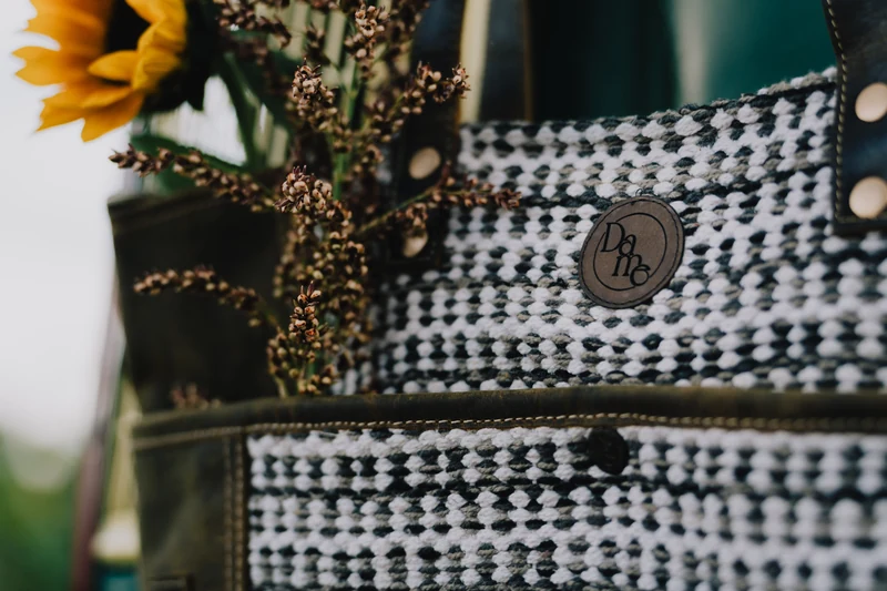 Close view of a Dame Renovations tote’s black-and-white weave, round brand patch, olive pocket, and sunflower with dried stems.