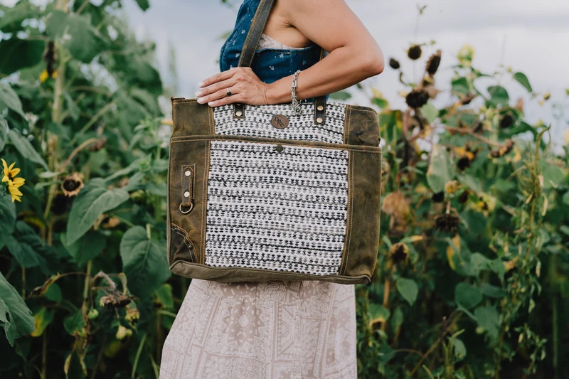 A person carries an olive tote with a black-and-white woven panel and brown handles among tall sunflowers.