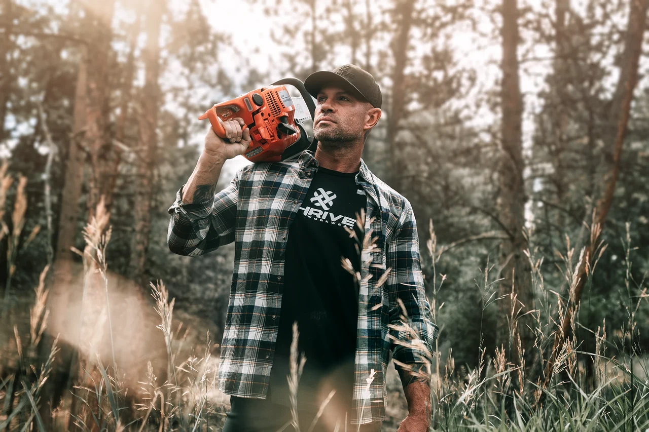 A man in a cap and plaid overshirt carries an orange chainsaw on his shoulder in a wooded setting, wearing a black THRIVE shirt.