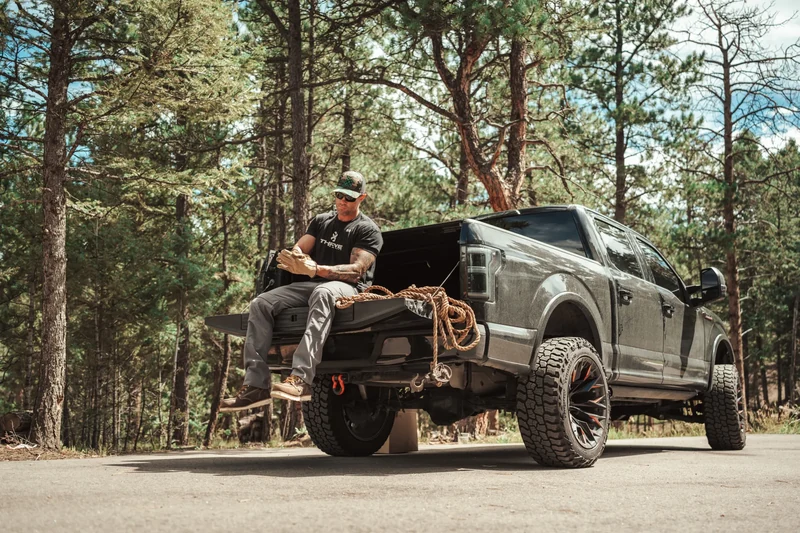 A man wearing a black shirt and work pants sits on a pickup truck’s tailgate beside coiled rope on a wooded roadside.