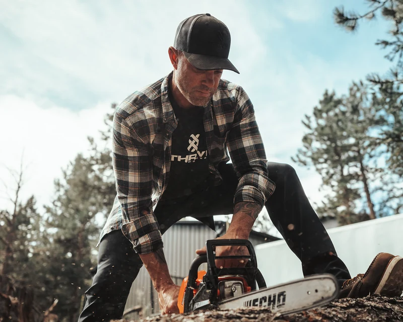 A man in a plaid overshirt and black THRIVE shirt uses a chainsaw to cut a log outdoors.